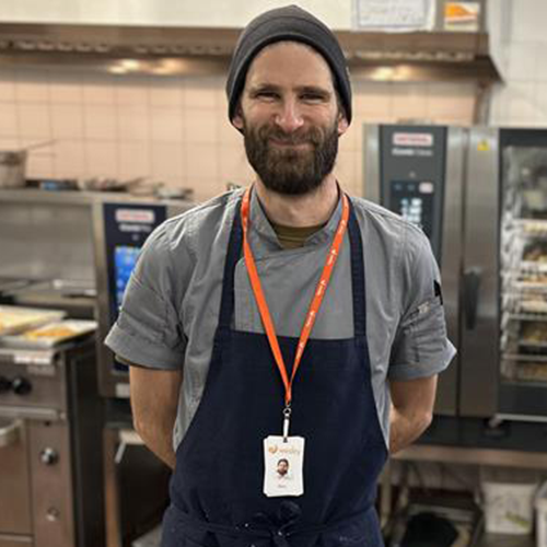 Alan in a commercial kitchen. He is smiling at the camera wearing an apron, beanie, and staff lanyard, with kitchen equipment behind him.