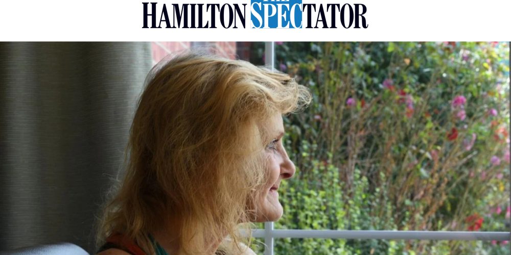 A woman with blonde hair sits indoors, smiling and looking out a window. Sunlight illuminates her face, and a garden with green plants and pink flowers is visible outside. The Hamilton Spectator logo at the top of the image.