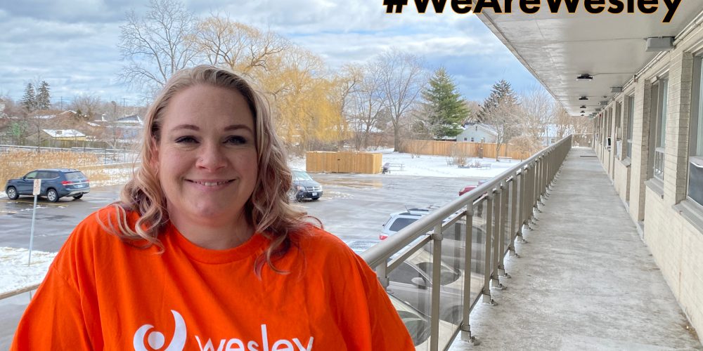 A woman in an orange wesley shirt stands on an outdoor walkway. Snow and parked cars are visible in the background.
