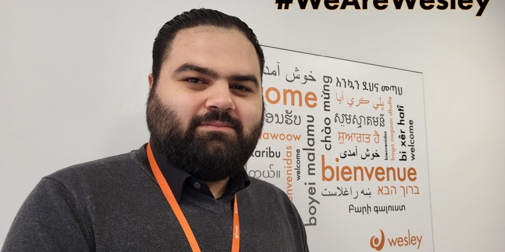 A man with a beard wearing an orange Wesley lanyard stands in front of a multilingual welcome sign.
